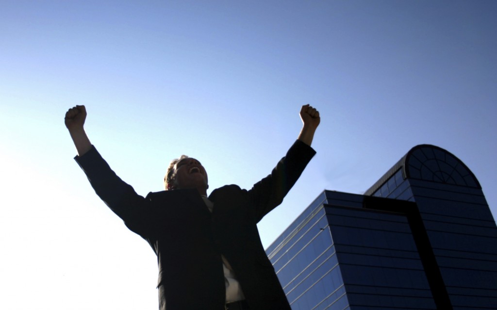 Business man holding up his arms in victory with a blue office building in the background